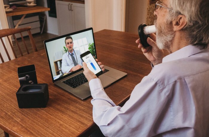 Man sitting at a table having a video call with a doctor, who is displayed on a laptop. The man is holding a phone, whilst blowing into a spirometer.